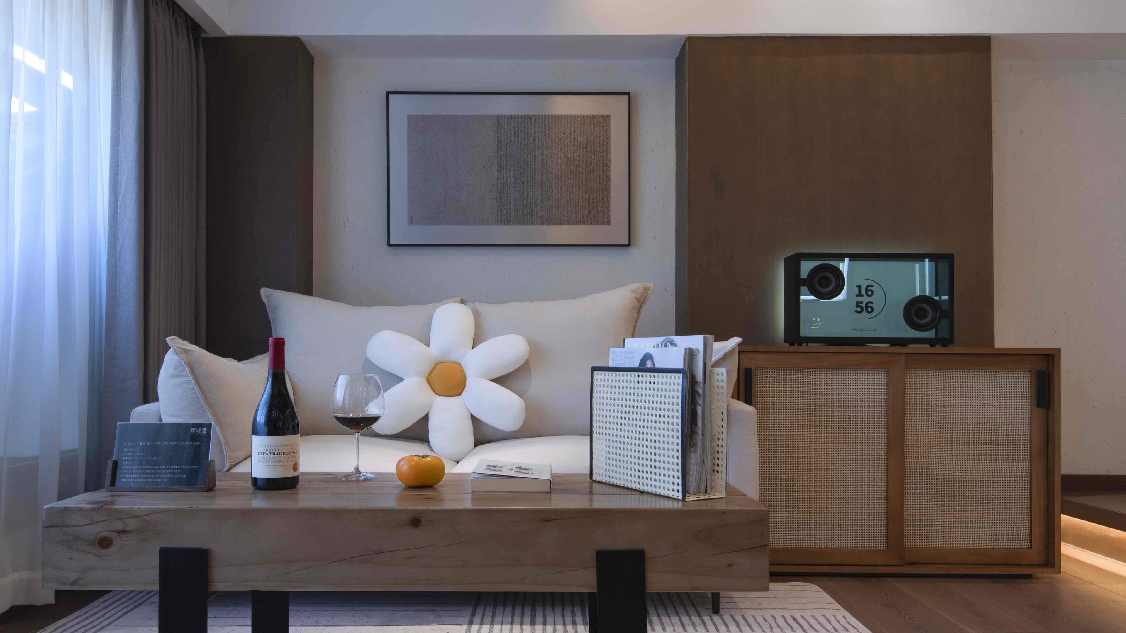 A modern living room with a wooden coffee table, a large flower-shaped pillow, a bottle of wine, a glass of wine, a small orange, and a framed picture on the wall.