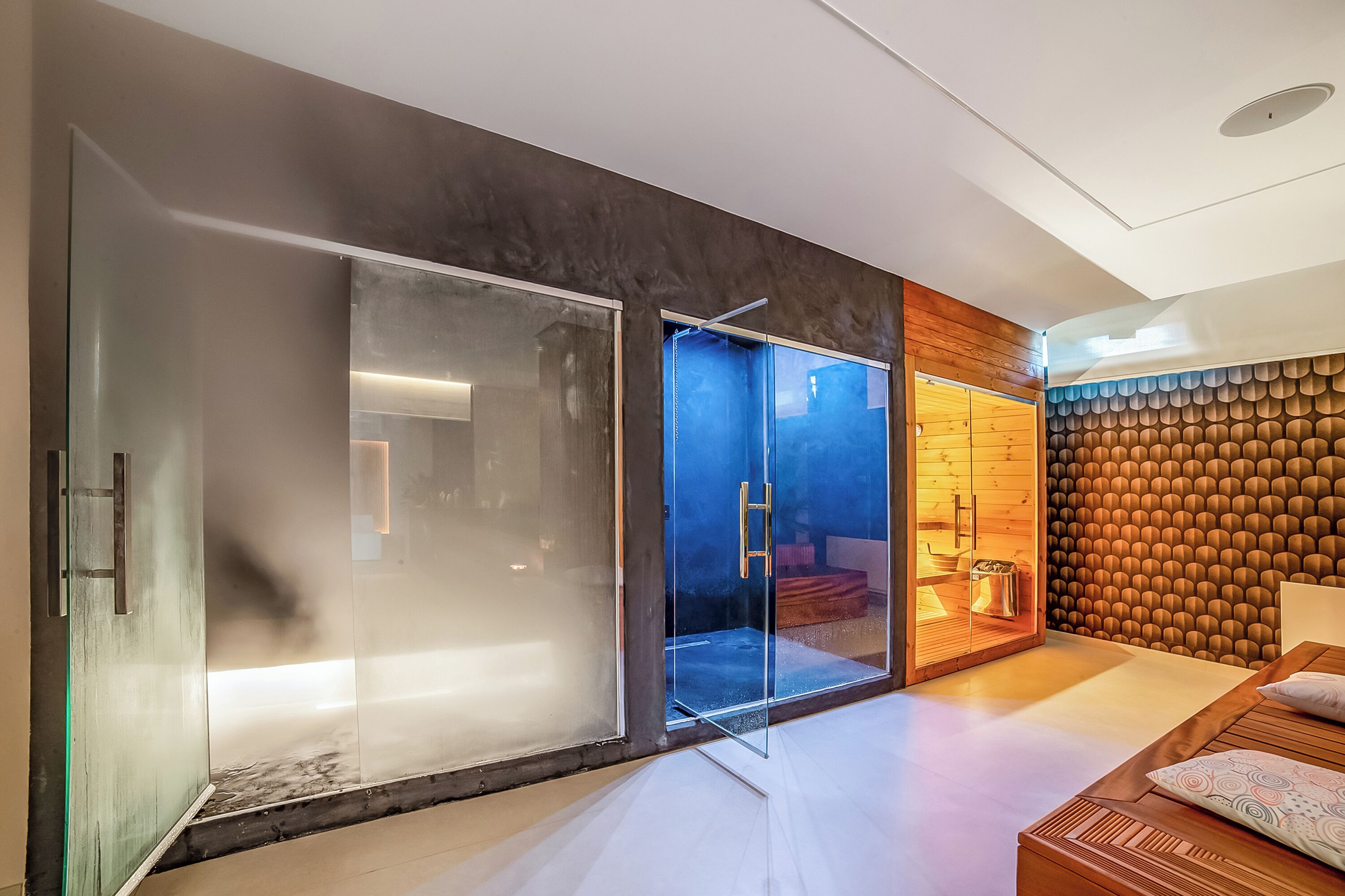 A modern interior with a glass door leading to a shower area, a wooden wall with a unique pattern, and a bed with a patterned pillow.