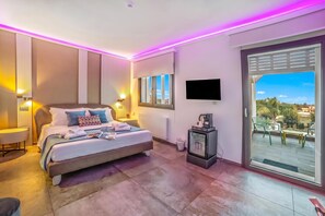 A modern hotel room with a large bed, a flat-screen TV, a balcony with outdoor seating, and a minibar.