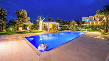 A swimming pool with a glass enclosure, surrounded by lounge chairs and umbrellas, and a well-lit patio area.