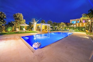 A swimming pool with a glass enclosure, surrounded by lounge chairs and umbrellas, and a well-lit patio area.
