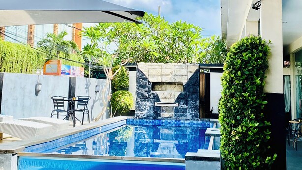 A swimming pool with steps, a stone wall, and a view of a building with large windows. - V Canggu Dormitory (Tibubeneng)