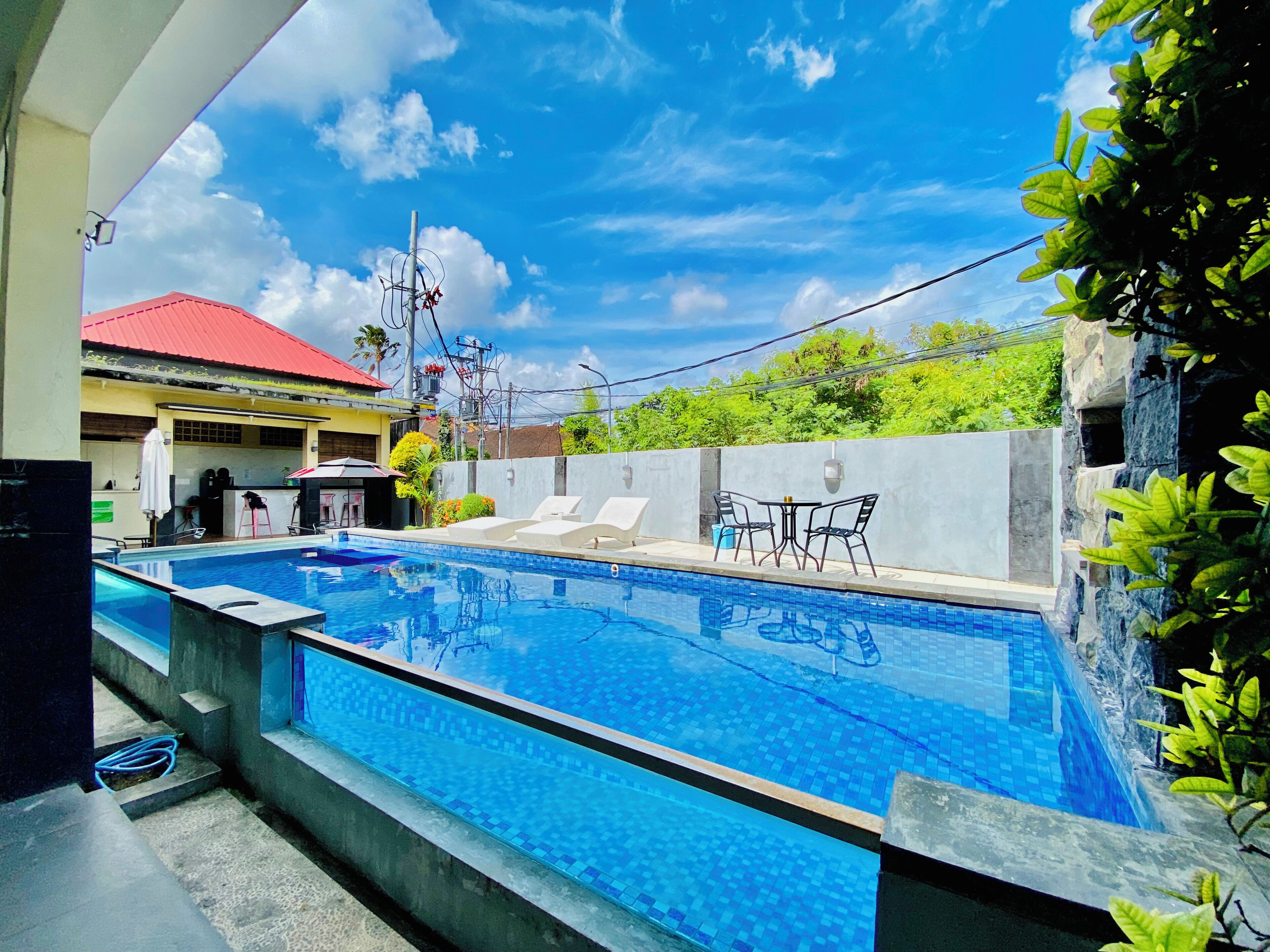 A swimming pool with lounge chairs and a table, surrounded by greenery and a building with a red roof.