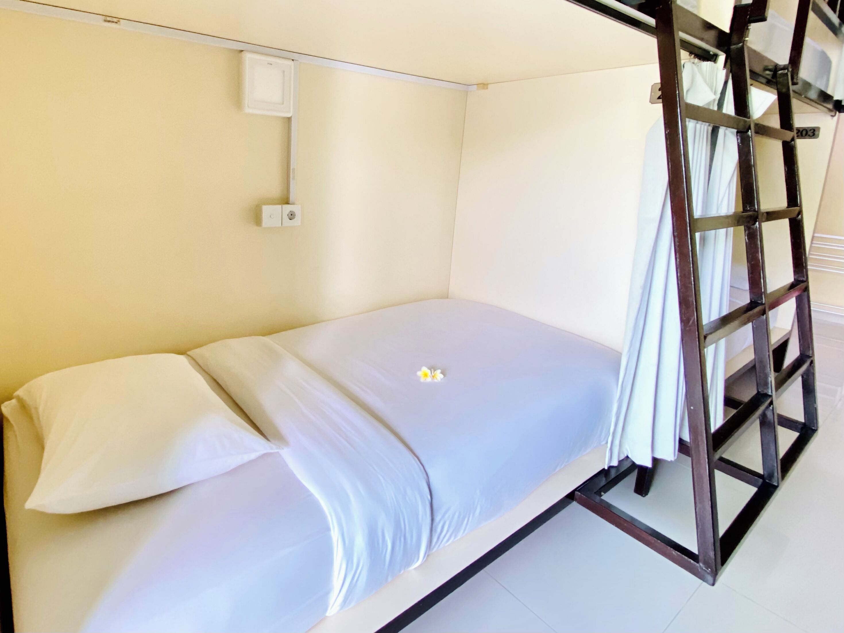 A single bed with a white mattress and pillows, a wooden ladder, a white wall, and a small electrical outlet.