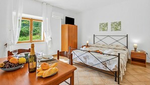Ein Schlafzimmer mit Bett, einem Holzschrank, einem Nachttisch mit Lampe, einem Fenster mit Vorhängen und einem Tisch mit Früchten und einer Flasche.