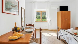Ein Zimmer mit Esstisch für zwei Personen, einem Bett mit Metallgestell, einem Holzschrank, einem fenster mit Vorhängen und einem gerahmten Bild an der Wand.