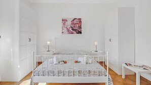 Een slaapkamer met een wit bed, twee nachtkastjes, een wandlamp en een schilderij aan de muur, plus een deur.