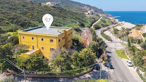 A yellow building with a rooftop terrace, surrounded by greenery and a coastal road. - Residence Argentiera Studio 12 Standard, Miniera dell’Argentiera, Italy (Miniera dell’Argentiera)
