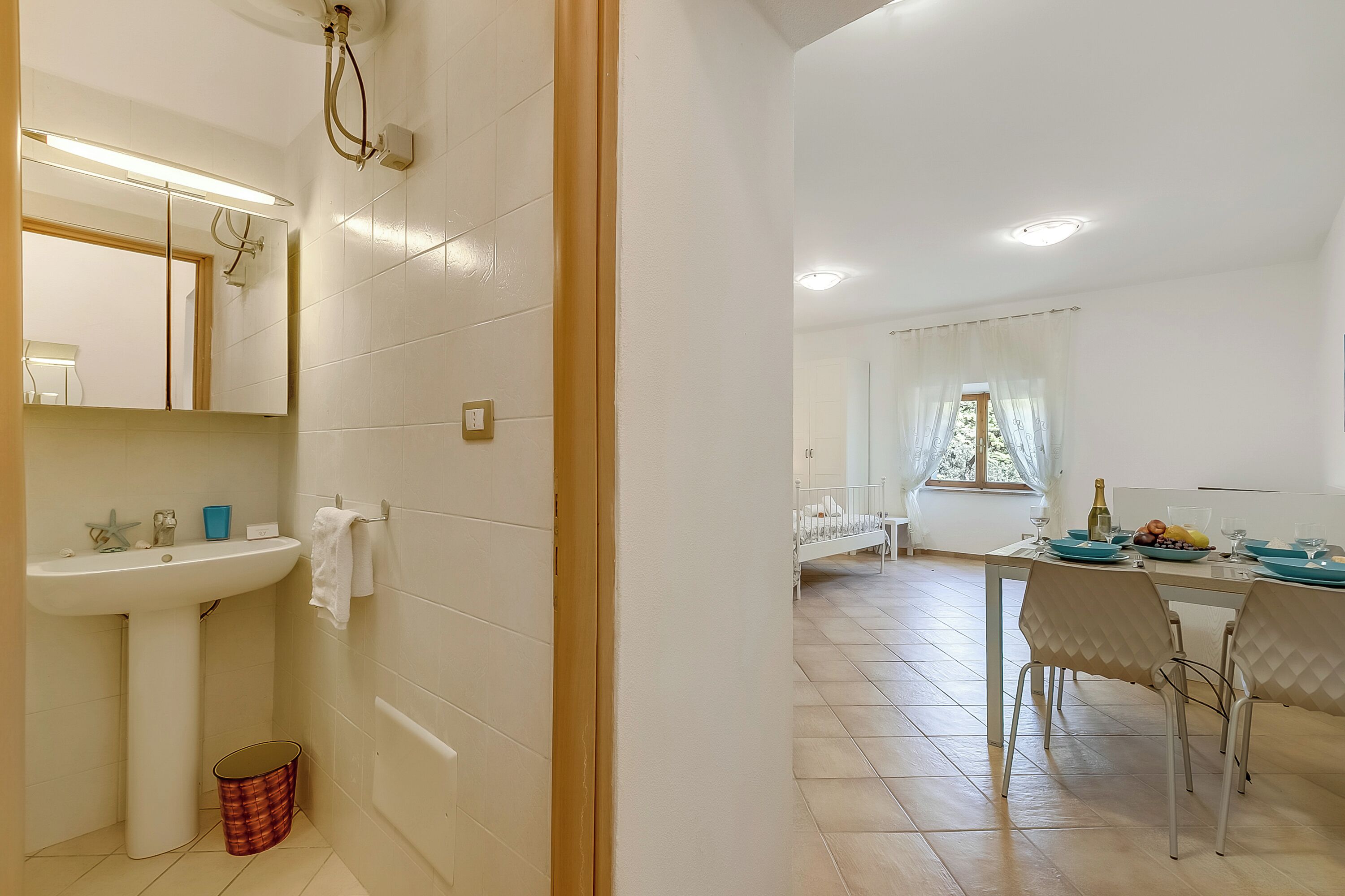 Une salle de bain équipée d’un lavabo, d’un miroir et d’un porte-serviettes. On aperçoit à l’arrière-plan un coin repas avec une table, des chaises et une fenêtre.