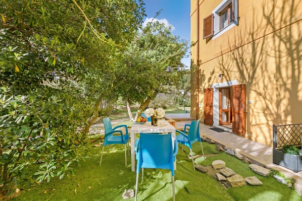 A patio with a table and chairs, surrounded by greenery and a building.