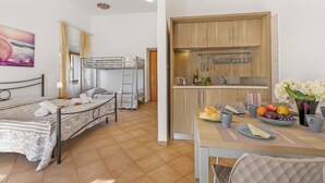 A room with two beds, a kitchen with wooden cabinets, a dining table with fruit and cups, and a painting on the wall. - Residence Argentiera Studio 10 for four, Miniera dell’Argentiera, Italy (Miniera dell’Argentiera)