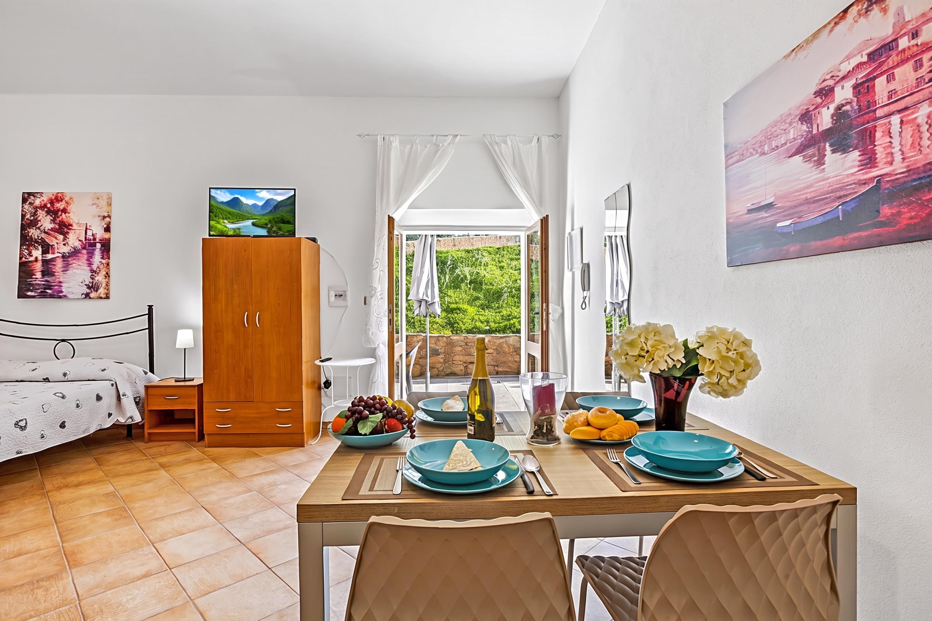 A hotel room with a bed, a wooden wardrobe, a dining table with blue plates, a bottle of wine, and a vase with flowers.