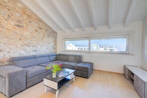 A modern living room with a stone wall, grey sectional sofa, a glass coffee table with a vase of flowers, and a view of buildings through a large window. - Peschiera Lake View + Parking, Peschiera del Garda, Italy (Peschiera del Garda)