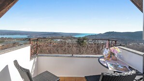 Ein Balkon mit Blick auf einen See und Hügel, ein Tisch mit einer Flasche und Blumen sowie Korbmöbel.