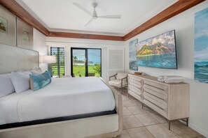 Ein Schlafzimmer mit Bett, Kommode, Sessel, Deckenventilator und einem großen Fenster mit Blick ins Freie.