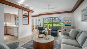 A modern living room with a round coffee table, grey sofas, a TV, and a view of a tropical landscape through large windows. - Poipu Sands Unit 514 Oceanfront 2 Bedroom 2 Bath (Poipu)