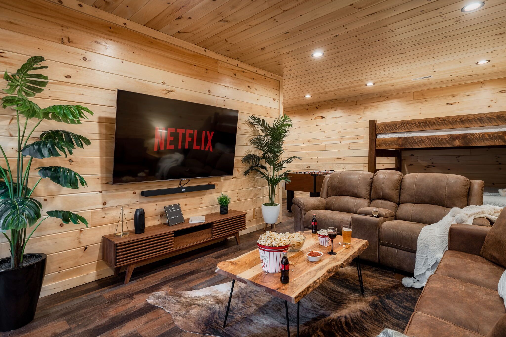 Ein gemütliches Wohnzimmer mit einem großen Flachbildfernseher, bequemen Ledersofas, einem Holztisch mit Snacks und einer Topfpflanze.