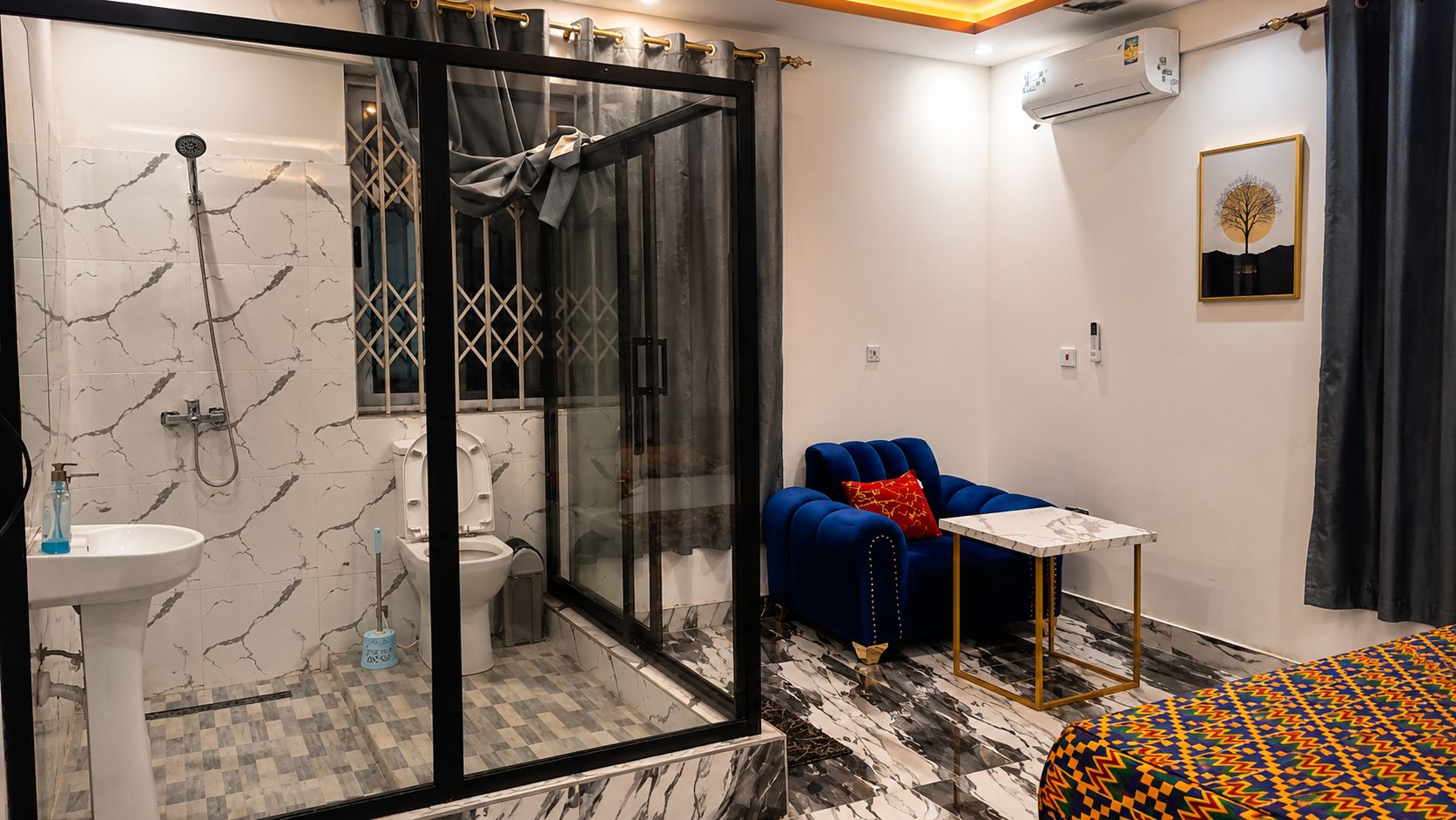 A modern bathroom with a glass shower enclosure, a white sink, and a toilet. There is a blue armchair with a red pillow and a small white table with a marble top.
