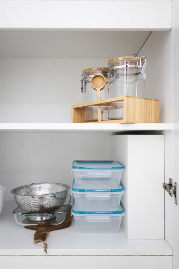 A white cabinet with two shelves. The top shelf holds two glass jars with metal lids and a wooden spoon. The bottom shelf has three clear plastic containers with blue lids and a metal strainer.