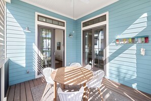 A covered patio with a wooden table and white chairs, glass doors, and a wall-mounted light fixture. - Sage & Sand: Golf Cart, Bikes & Pickleball (Santa Rosa Beach)