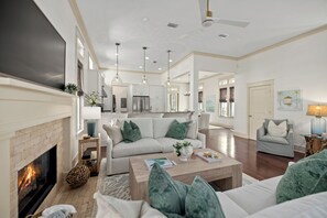 A spacious living room with a fireplace, a sofa set, a coffee table, and a kitchen in the background. - Sage & Sand: Golf Cart, Bikes & Pickleball (Santa Rosa Beach)