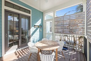 A covered patio with a round wooden table and white chairs, a glass door leading to an outdoor area, and a white fence with decorative vines. - Sage & Sand: Golf Cart, Bikes & Pickleball (Santa Rosa Beach)