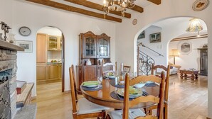 Ein Essbereich mit einem Holztisch, Stühlen und einer Chandelüster. Durch einen Bogen ist eine Küche zu sehen, und eine Treppe führt zu einer anderen Ebene.