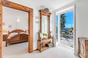 A bedroom with a wooden bed, a large mirror, and a dresser. There is a balcony with a view of the mountains. - Casa Naima: Family House | Terrace & Peaceful Location, Fescoggia (Alto Malcantone), Switzerland (Fescoggia (Alto Malcantone))