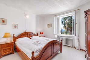 Ein Schlafzimmer mit einem hölzernen Bett, Nachttischen, einem Fenster mit Blick auf Palmen und einer Heizung.