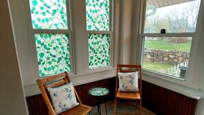 Une véranda avec trois fenêtres ornées de motifs de feuilles, deux fauteuils en bois avec des coussins à motifs floraux et une petite table ronde.