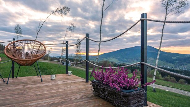 A wooden deck with a wicker chair, a table with two cups, and a basket of purple flowers. - Lovely home in Niedzwiedz with sauna (Niedzwiedz)