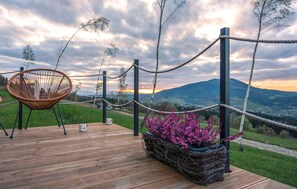 Una terraza de madera con una silla de mimbre, una mesa con dos tazas y una cesta de flores moradas.