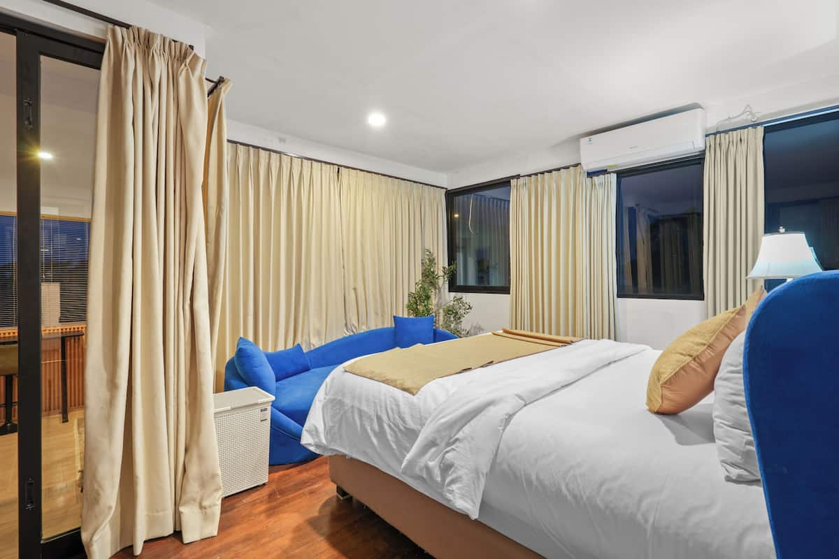 A hotel room with a bed, a blue sofa, a window, curtains, and an air conditioning unit.