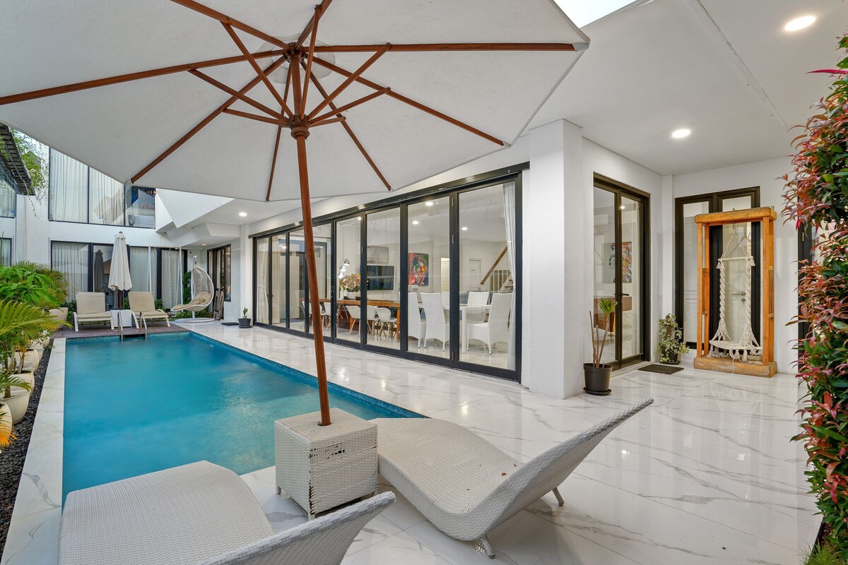 Un área de piscina interior moderna con sillones reclinables, una sombrilla grande y un comedor con paredes de vidrio.