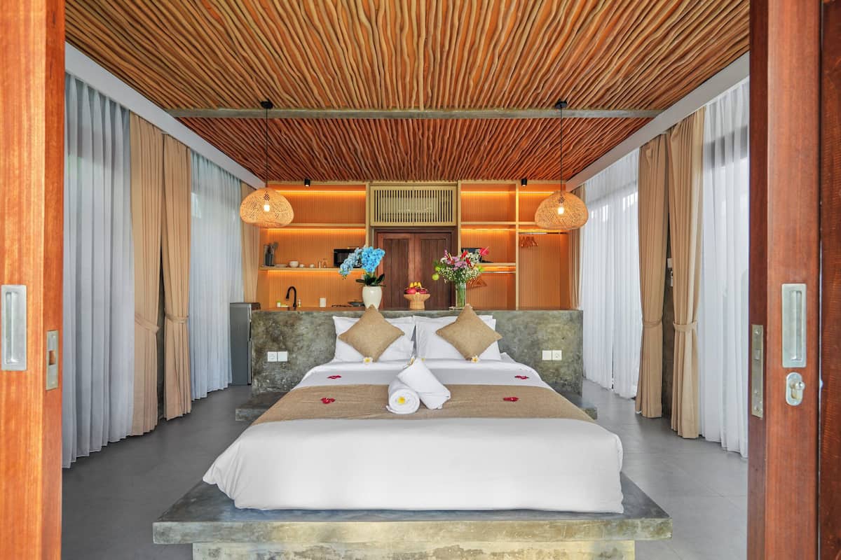 A hotel room with a bed, wooden ceiling, curtains, and a reception area in the background.