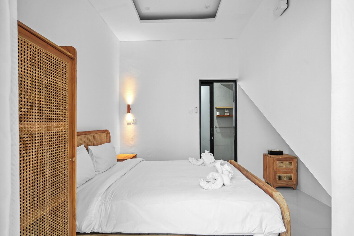 Una habitación de hotel moderna con cama, cabecero de madera, luz de pared, mesita de noche de madera y armario con puerta de mimbre.