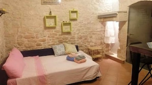 A cozy room with a stone wall, a bed with pillows, a small table, and a window with curtains. - Studio directly in the historic center (Giovinazzo)