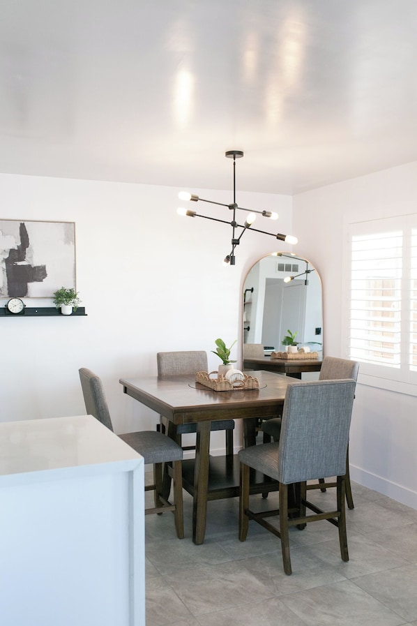 A dining area with a wooden table, grey upholstered chairs, a black chandelier, a round mirror, and a wall-mounted clock.