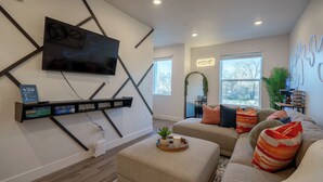 A modern living room with a flat-screen TV mounted on the wall, a sectional sofa, ottomans, and a decorative wall piece. - Modern Townhome 10 min to Downtown + Airport (Salt Lake City)