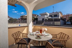 Un balcon avec une table ronde pour deux personnes, agrémenté de chaises en osier, d’un vase rempli de fleurs et d’une assiette de pâtisseries.