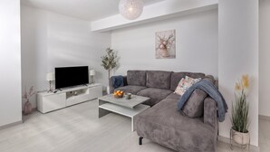 Een moderne woonkamer met een grijze bank, een flatscreen televisie op een witte kast, een salontafel en een hanglamp.