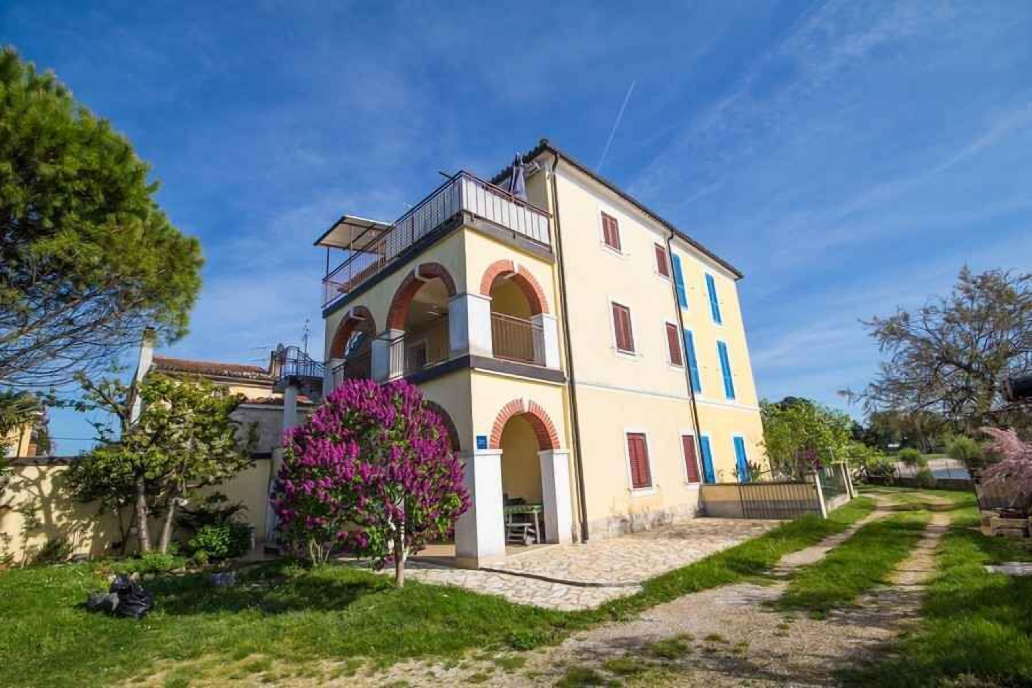 Un bâtiment à deux étages doté d’un balcon, de portes en arc de cercle et d’un jardin avec un arbre.