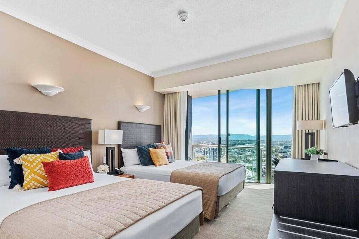 A hotel room with two beds, a desk, a TV, and a large window with a city view.