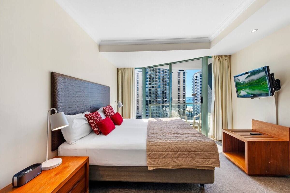 A hotel room with a bed, a TV, a desk, and a view of tall buildings.