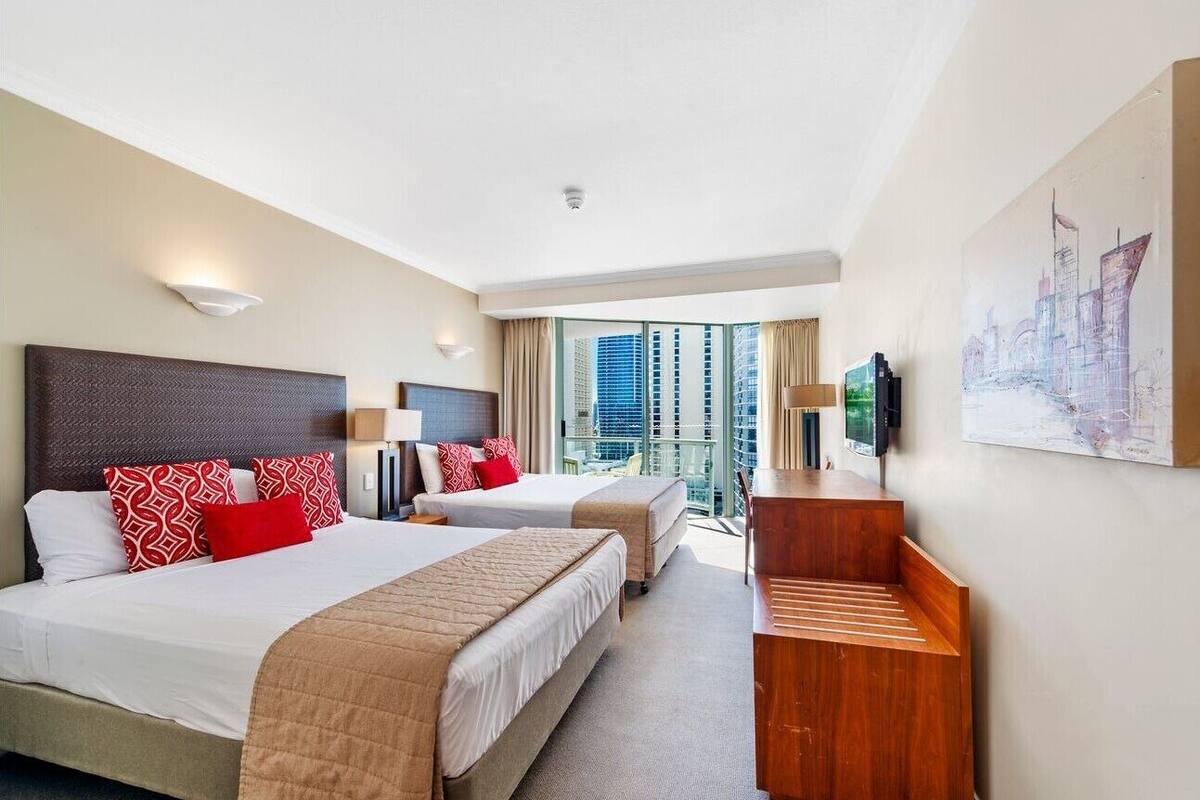 A hotel room with two beds, a desk, a TV, and a balcony with a city view.