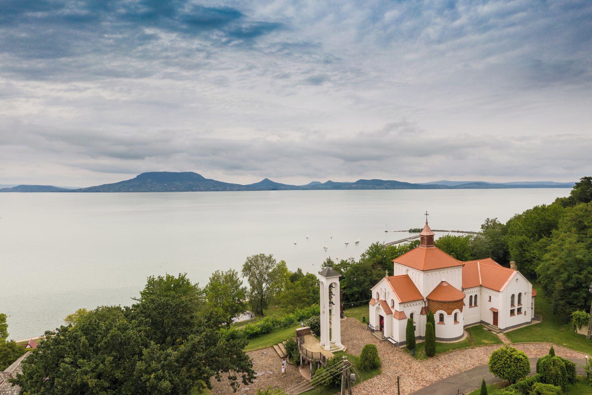 A church with a bell tower and a red roof, surrounded by greenery and a lake.