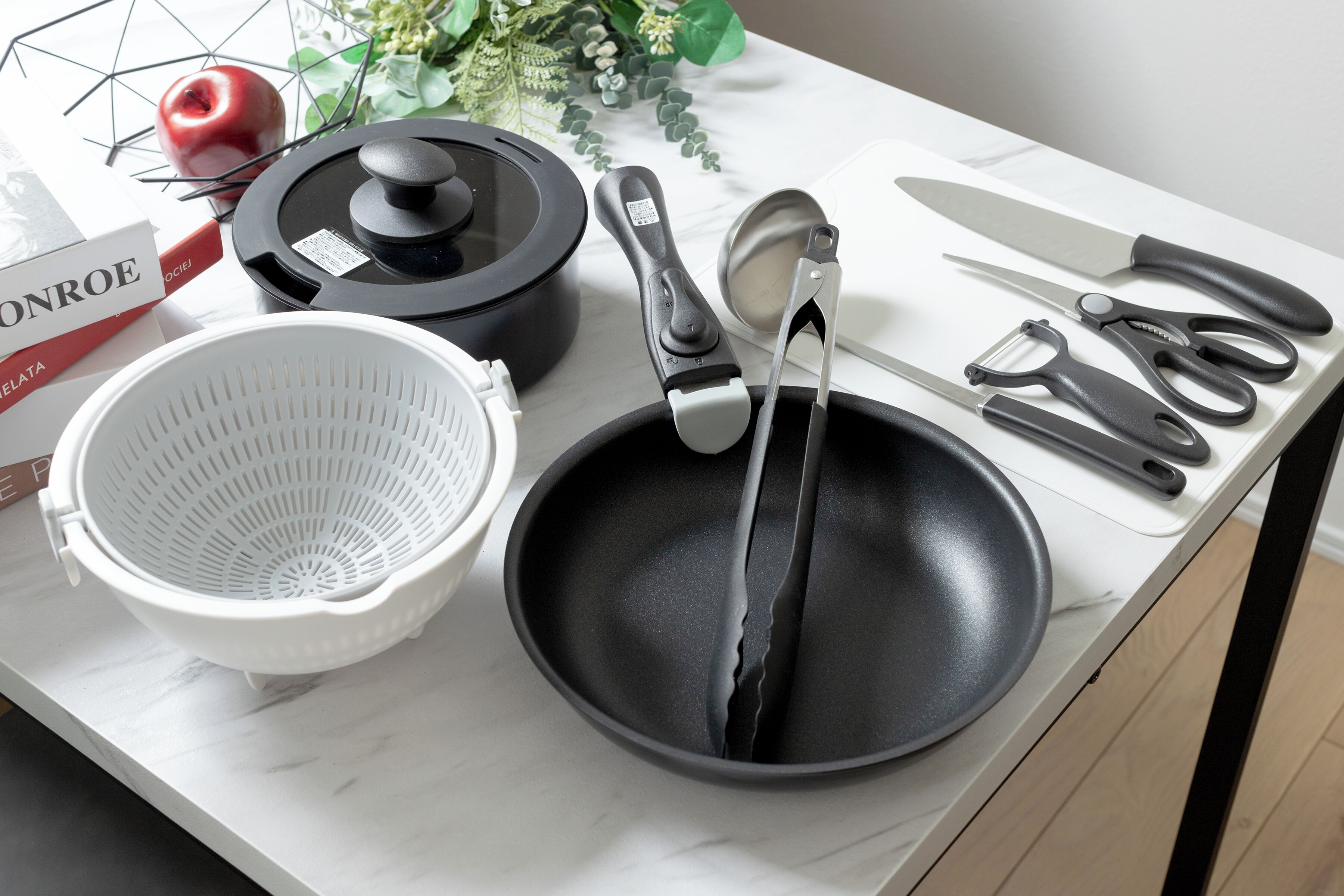 Una encimera de cocina con una sartén negra, un colador blanco, una olla negra, un par de tijeras, un cuchillo y una manzana roja.