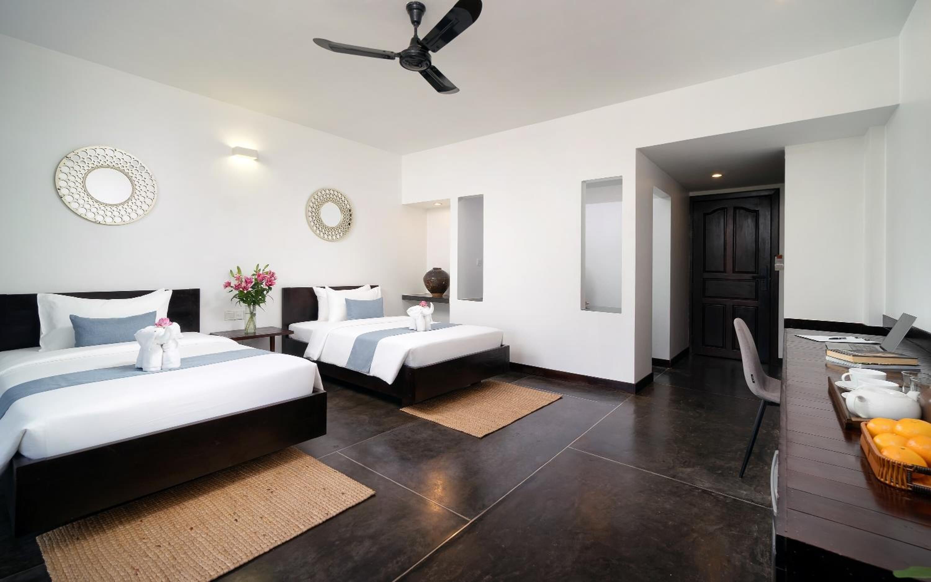 A hotel room with two beds, a ceiling fan, a desk with a laptop, and a door leading to another room.