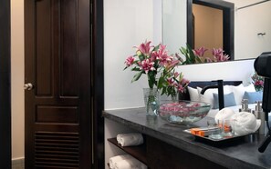 Un baño con puerta de madera oscura, un espejo, un lavabo, un cuenco de vidrio con flores rosadas, una toalla y artículos de aseo.