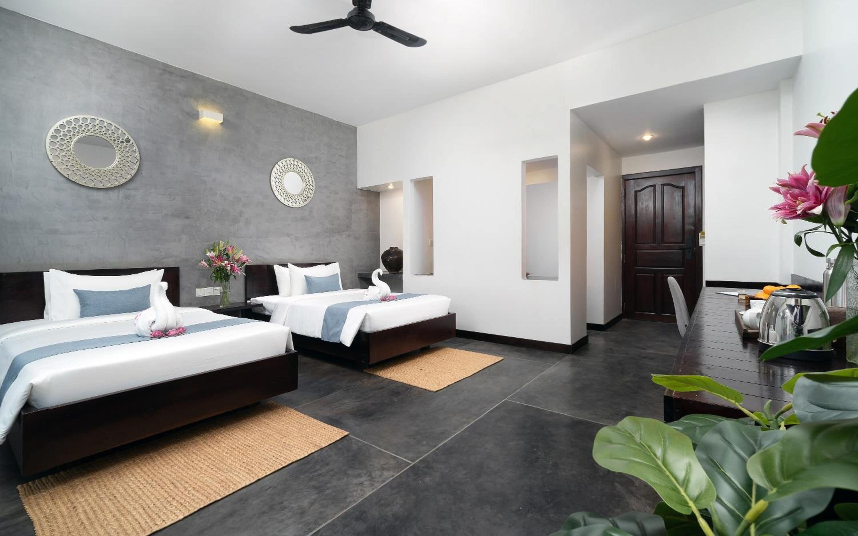 A modern hotel room with two beds, a ceiling fan, and a view of the hallway.